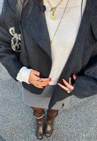 Blazer grigio di lana sopra a un maglione chiaro, gonna grigia e stivali di pelle marrone. Collane a strati in oro e argento, anelli e smalto per unghie.