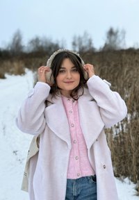Jeune femme ajustant des cache-oreilles beiges, portant un manteau rose clair et un pull, debout sur un sentier enneigé avec de l'herbe sèche dans un paysage d'hiver.