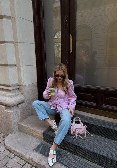 Mujer con gafas de sol, blusa rosa y jeans sentada en escalones de piedra frente a un edificio, sosteniendo una bebida verde con un bolso rosa cerca.