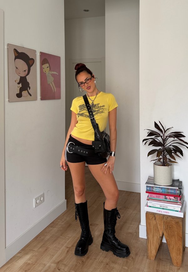 Jeune femme posant à l'intérieur portant un t-shirt jaune, un short noir, des bottes noires, un sac en bandoulière, avec deux peintures de dessins animés et une plante sur des livres à proximité.