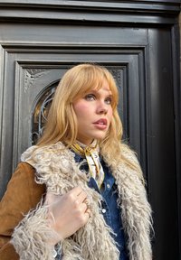Tan ruskindsjakke med en fluffy beige pelskrave, en blå denimskjorte under og et mønstrede gult og hvidt silkeslør.