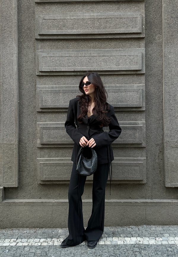 Mujer con gafas de sol negras y traje negro a medida sosteniendo un bolso negro texturizado, de pie frente a una pared gris texturizada.