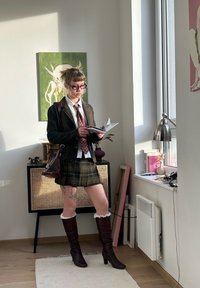 Ung person i rutet skjørt og brun jakke, leser en bok. Merkbare brune knelange støvler og briller; moderne interiørdekor synlig.