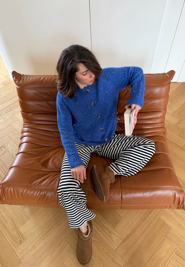 Person in einem blauen Pullover und gestreiften Hosen sitzt im Schneidersitz auf einem braunen Ledersofa, hält ein Buch und schaut nach unten.