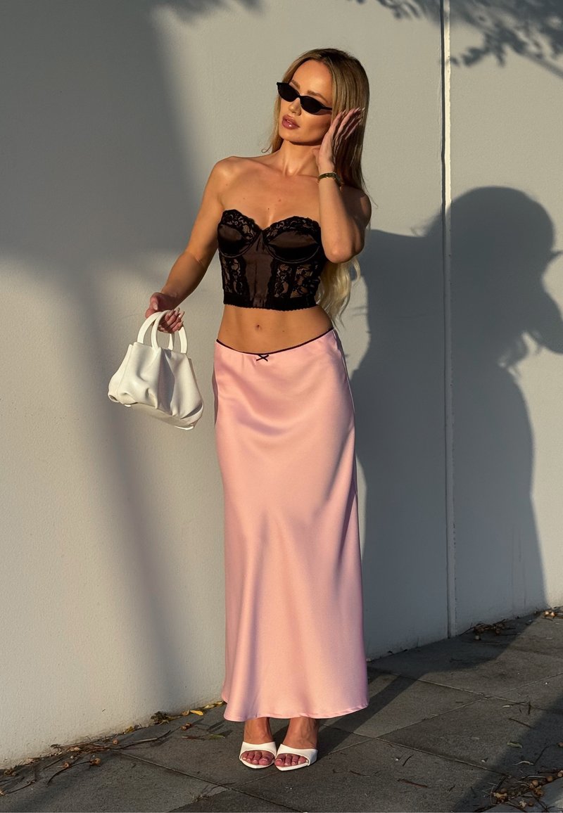 Femme aux longs cheveux portant un bustier en dentelle noire, une jupe en satin rose, des talons blancs, des lunettes de soleil noires, tenant un sac à main blanc contre un mur ensoleillé.