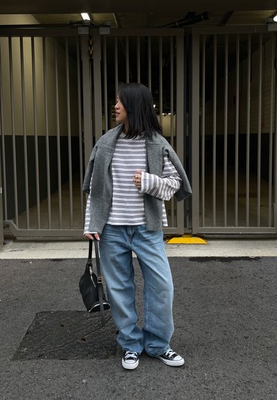Una persona está de pie en un estacionamiento, vistiendo un suéter gris, una camisa a rayas, jeans azules holgados y zapatillas negras, y lleva una bolsa negra.