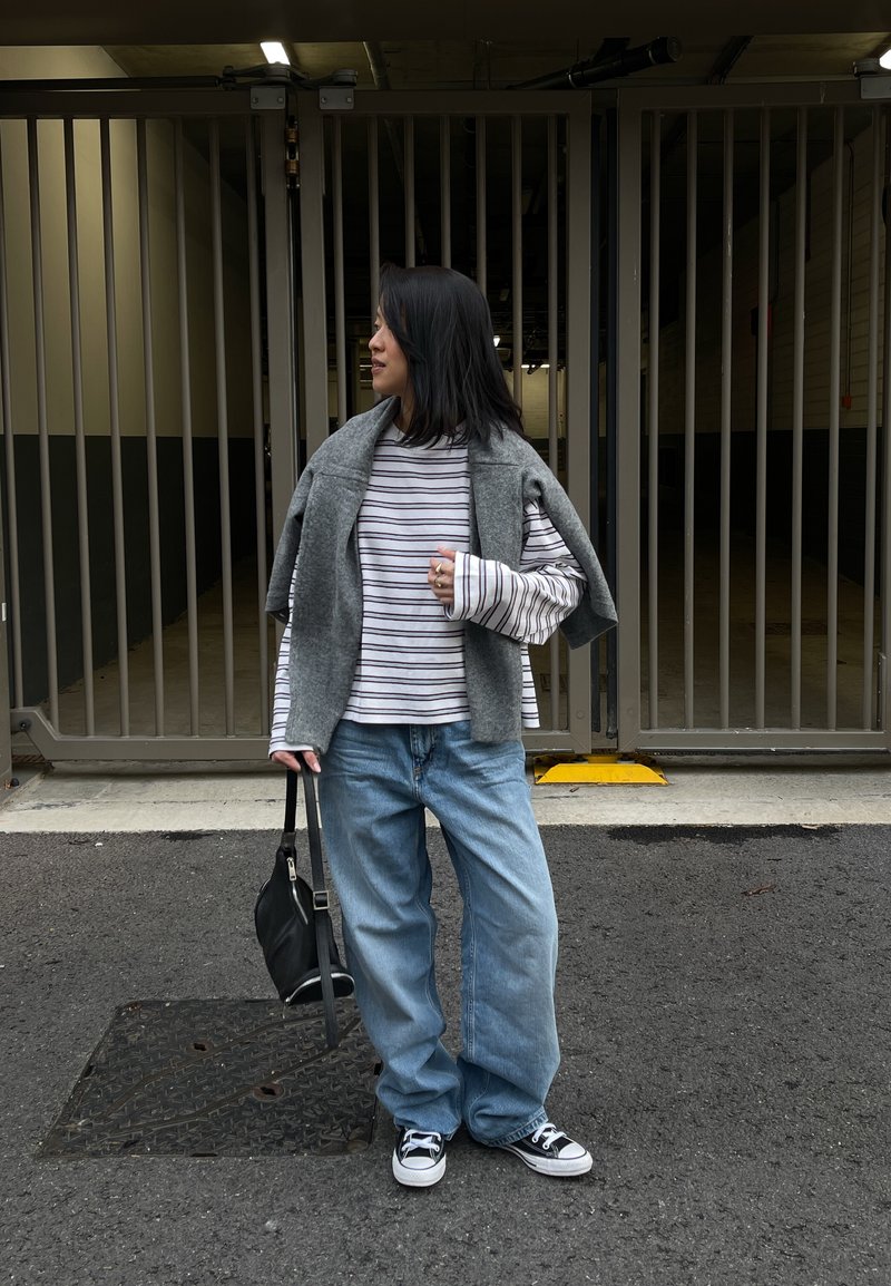Une personne se tient dans un parking en portant un pull gris, une chemise rayée, un jean bleu ample et des baskets noires, tenant un sac noir.