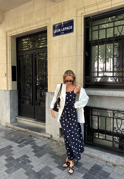 Mujer con un vestido negro de lunares y una blazer blanca se encuentra en la acera de la ciudad cerca de un edificio beige con puertas de hierro forjado negro y un cartel de calle.