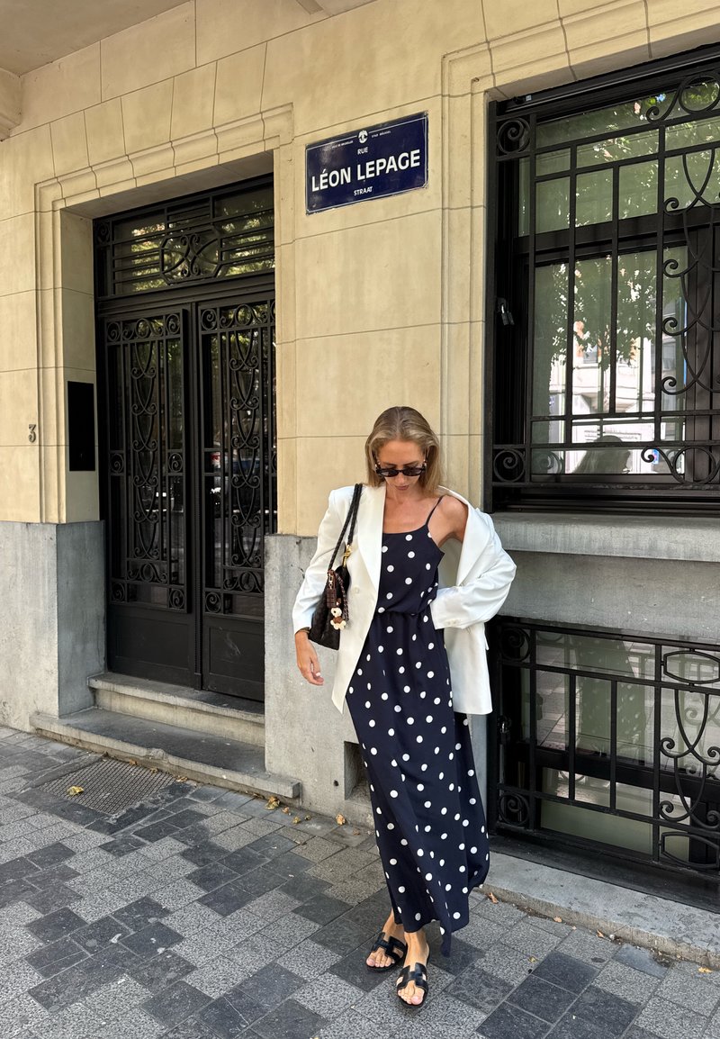 Donna in un vestito nero a pois e giacca bianca si trova sul marciapiede della città vicino a un edificio beige con porte in ferro battuto nere e un cartello stradale.