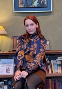 Jeune femme aux cheveux roux portant un chemisier à motifs bleu marine et orange, assise sur une chaise devant une bibliothèque et des cadres photo.