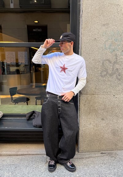 Joven con gorra negra, camisa blanca de manga larga con estrella roja, pantalones anchos negros y zapatos negros, de pie frente a la ventana de una cafetería.