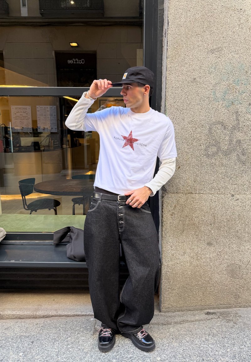 Ung mand iført sort kasket, hvid langærmet skjorte med rød stjerne, vidt sort bukser og sorte sko, der står udenfor et cafévindue.