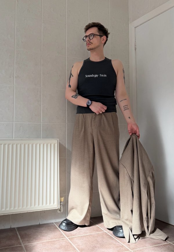 Person med briller og en svart ermelaus topp med tekst, holder en brun jakke, står mot en flislagt vegg nær en radiator.