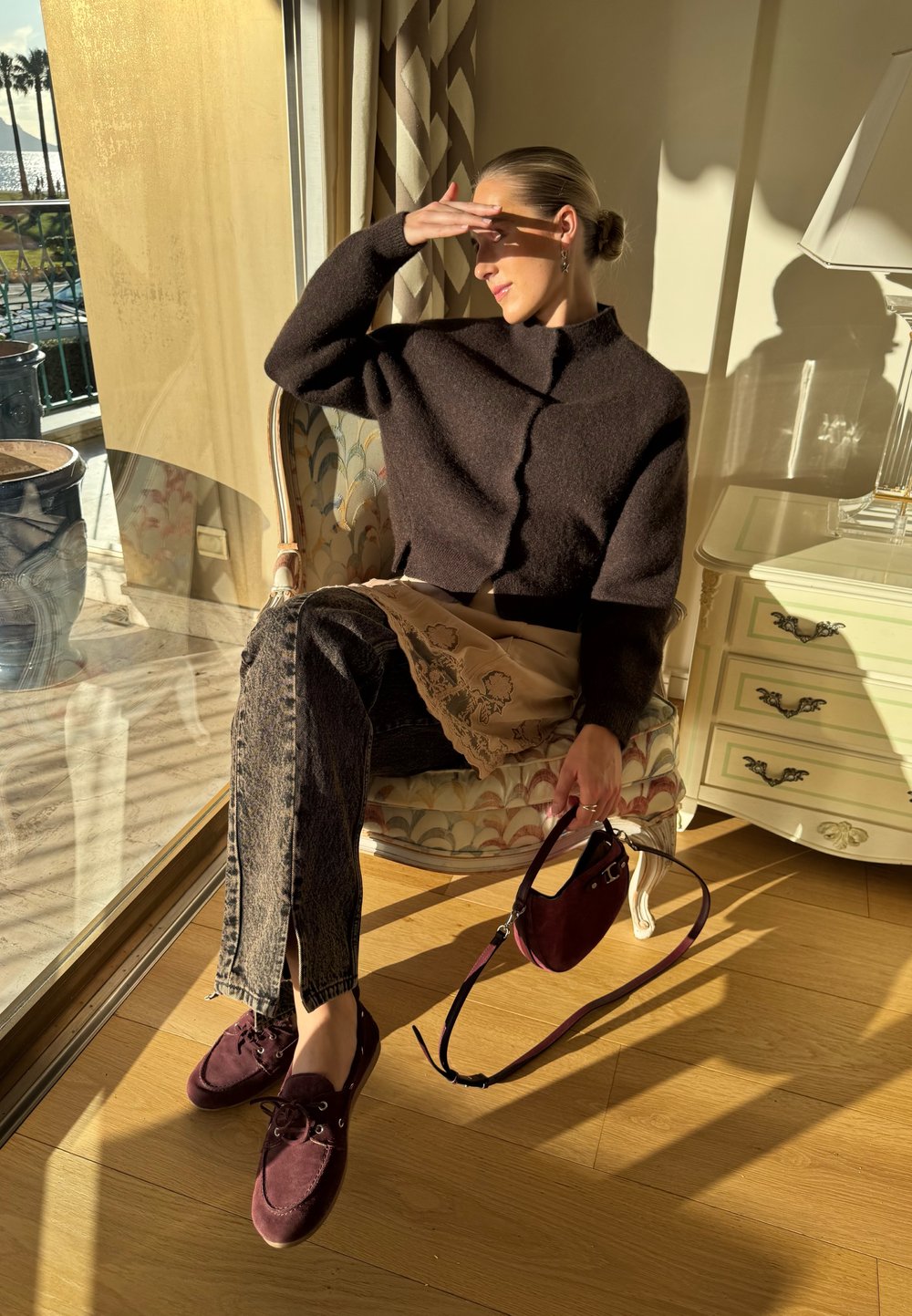 Femme assise sur un fauteuil à motifs près de la fenêtre, se protégeant les yeux du soleil, portant un pull sombre, un jean, des chaussures bordeaux et tenant un sac assorti.