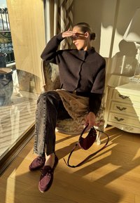 Femme assise sur un fauteuil à motifs près de la fenêtre, se protégeant les yeux du soleil, portant un pull sombre, un jean, des chaussures bordeaux et tenant un sac assorti.