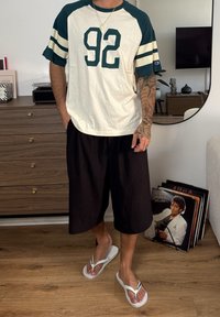 Hemd mit kurzen Ärmeln in Weiß und Dunkelgrün mit großem "92" Aufdruck, kombiniert mit schwarzen Shorts und weißen Flip-Flops. Tattoos sind am Arm sichtbar.