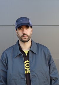 Chaqueta azul oscura con cremallera al frente, utilizada sobre una camisa negra con un patrón geométrico amarillo y un cuello amarillo. Gorra con acentos de logo.