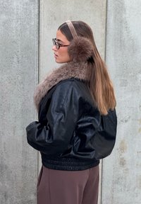 Casaco bomber de couro preto com gola de pelo sintético. Acompanhado por protetores auriculares de pelo castanho e uma faixa de cabeça macia bege. Textura lisa e corte descontraído.