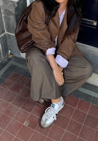 Brun læderjakke over en lyseblå-stribet skjorte, parret med løse brune bukser og hvide sneakers. Brun vævet taske hviler ved siden af.