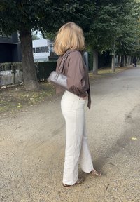 Ruskea ylipitkä takki, kermaväriset leveät housut ja leopardikuosiset kengät; pitää pientä harmaata käsilaukkua; hiukset kevyesti laineilla, keskikokoiset.