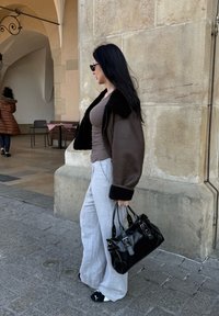 Jaqueta bombardeiro castanha com colarinho de pele preta, calças cinzentas de perna larga e uma bolsa de couro preta. Texturas suaves com um visual casual.