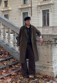 Long brown check-patterned coat over a blue argyle jumper, beige shirt, and dark trousers. Black cap. Leaves scattered on the ground.