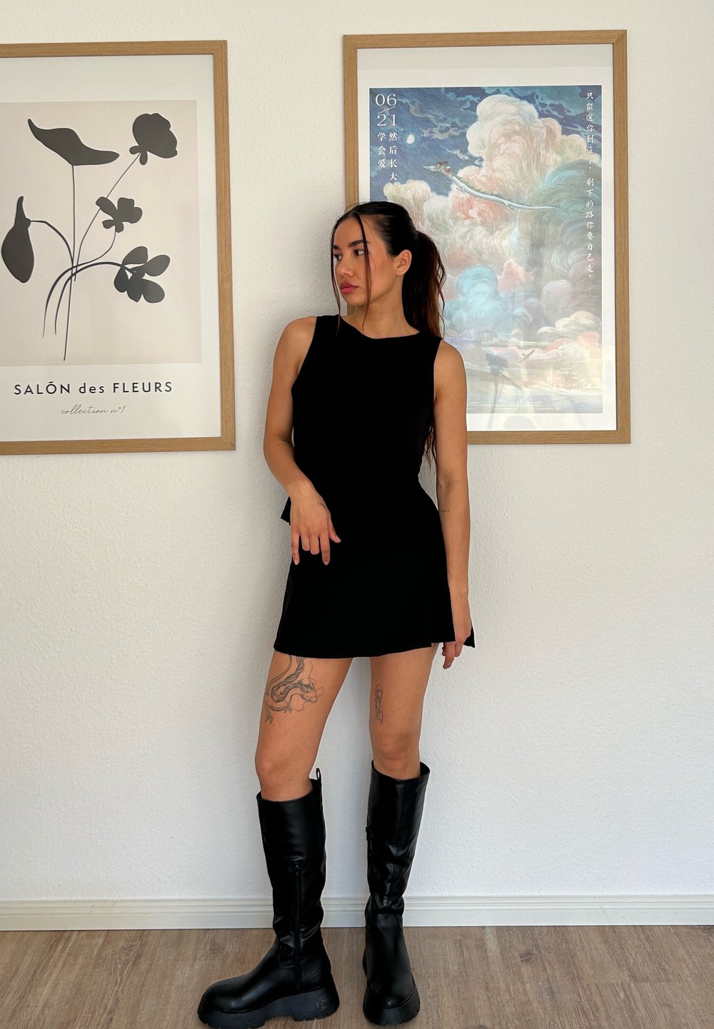 Čierny top bez rukávov v kombinácii s krátkou sukňou rovnakého odtieňa. Modelka má na sebe čierne vysoké čižmy. Svetlé drevené podlahy a rámované umelecké dielo v pozadí.