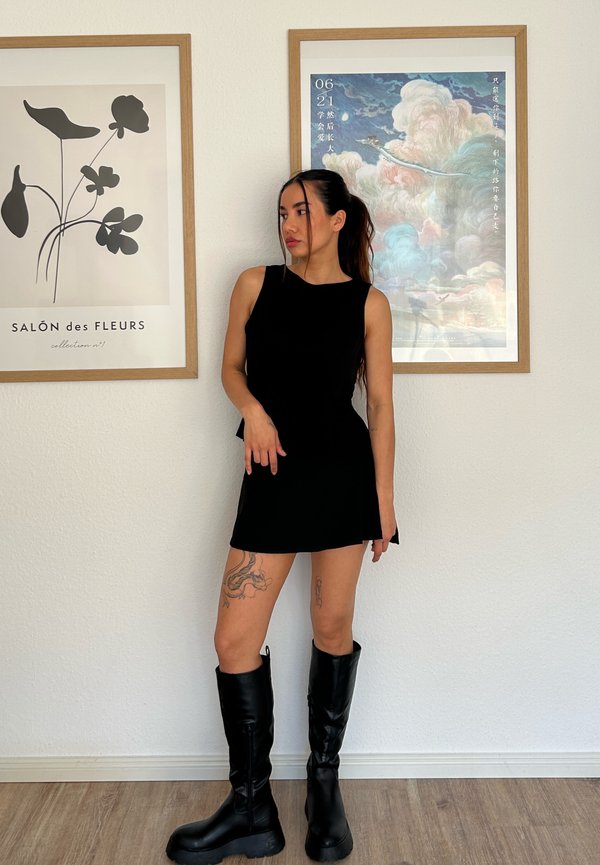 Top preto sem mangas combinado com uma saia curta a condizer. Modelo usa botas pretas até ao joelho. Chão de madeira clara e arte emoldurada ao fundo.