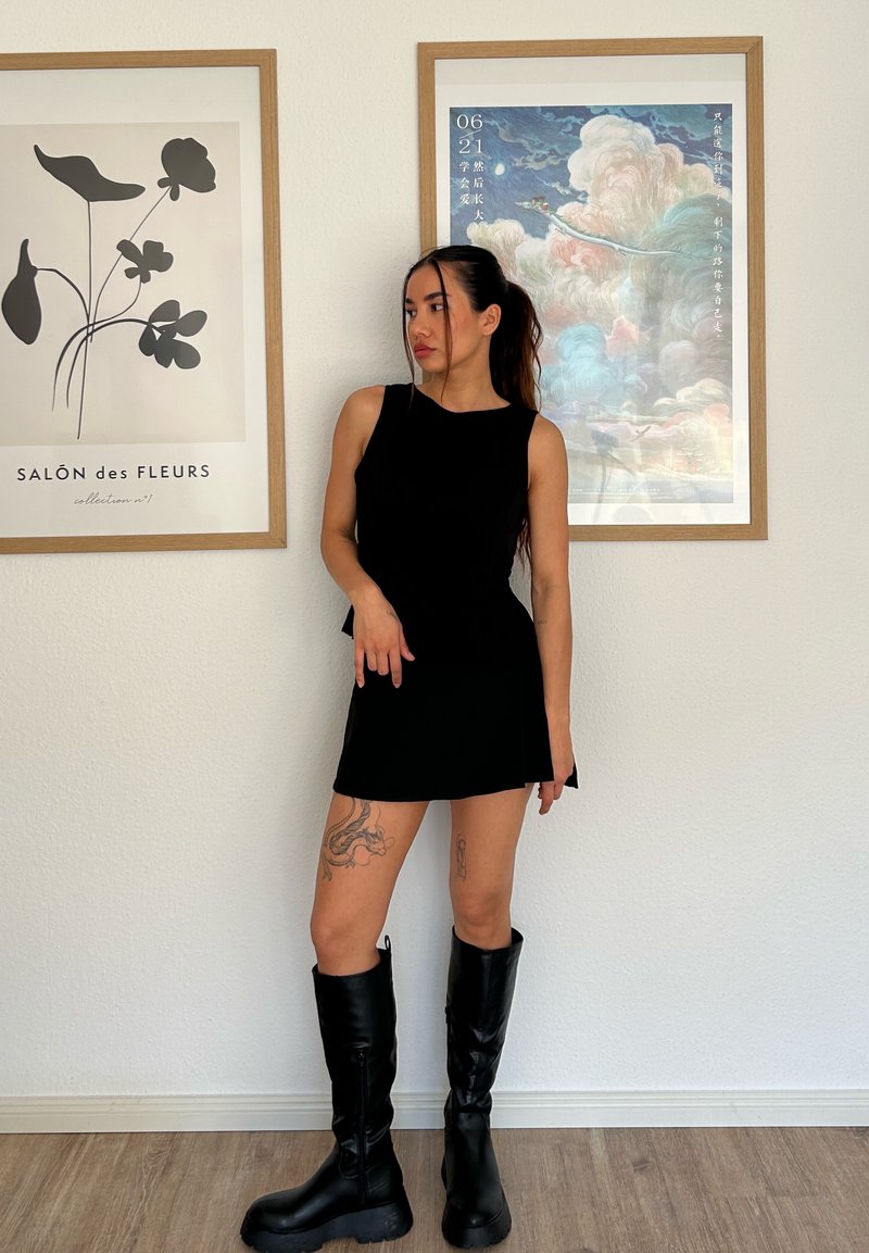 Top preto sem mangas combinado com uma saia curta a condizer. Modelo usa botas pretas até ao joelho. Chão de madeira clara e arte emoldurada ao fundo.
