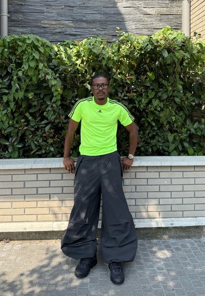 Camiseta atlética de manga corta en verde neón con tres rayas negras, combinada con pantalones negros de gran tamaño y zapatillas negras, con un fondo de vegetación.