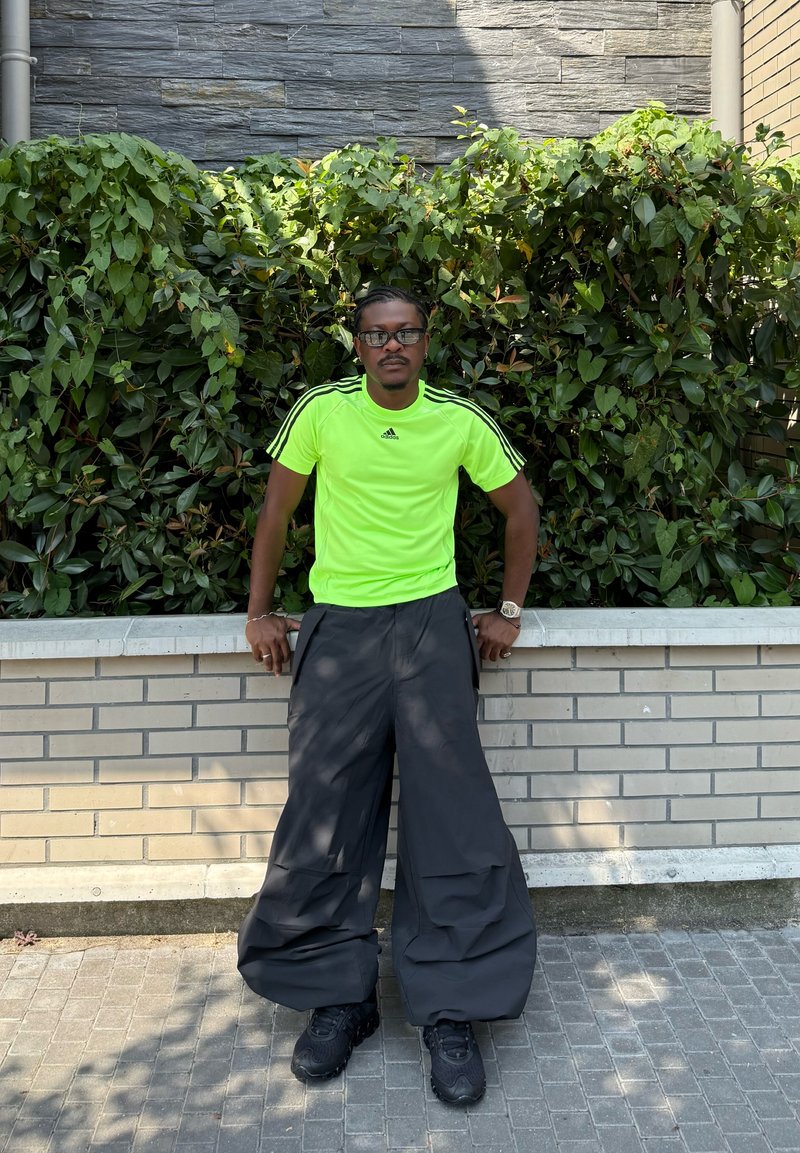 T-shirt de sport à manches courtes vert néon avec trois rayures noires, associé à un pantalon noir surdimensionné et des baskets noires, le tout sur un fond verdoyant.