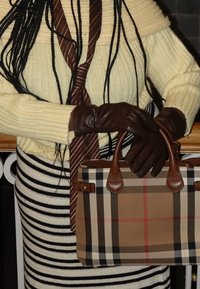 Personne portant un pull en tricot crème, une cravate rayée marron, une jupe rayée et des gants en cuir marron tenant un sac à main à carreaux beige.