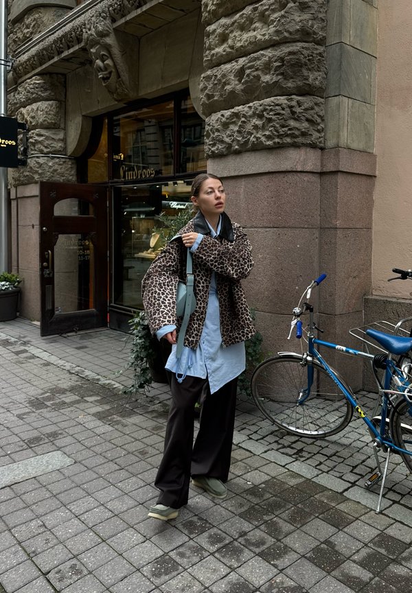 Jagthunde-jakke med leopardprint, lysebluse, sorte bukser, grønne sko. Sølvrygsæk, urban indstilling med brosten og cykel.