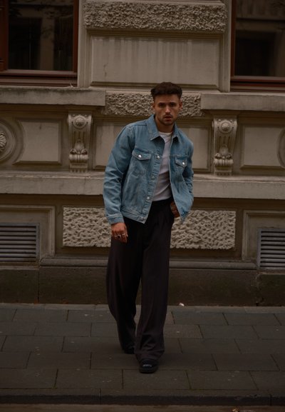 Hombre caminando por la acera de la ciudad, vestido con una chaqueta de mezclilla azul, camisa blanca y pantalones negros holgados, con las manos en los bolsillos, cerca de una fachada de edificio ornamentada.