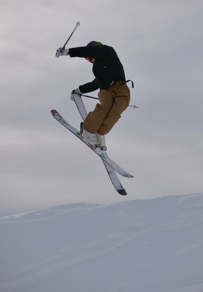 Esquiador con pantalones marrones y chaqueta negra realiza un salto en el aire sobre una pendiente nevada bajo un cielo nublado.