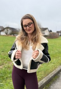 Zwart en crème faux shearling jack met brede kraag, wit gebreid topje en paarse kabelgebreide leggings. Buitenomgeving met groen.