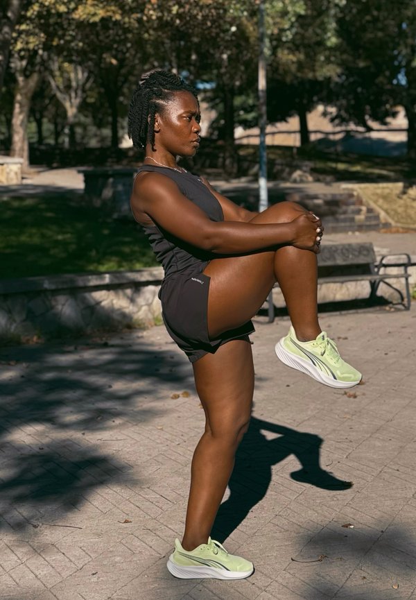 Femme en tenue de sport noire étirant une jambe en tirant le genou vers la poitrine en plein air par une journée ensoleillée dans un parc.