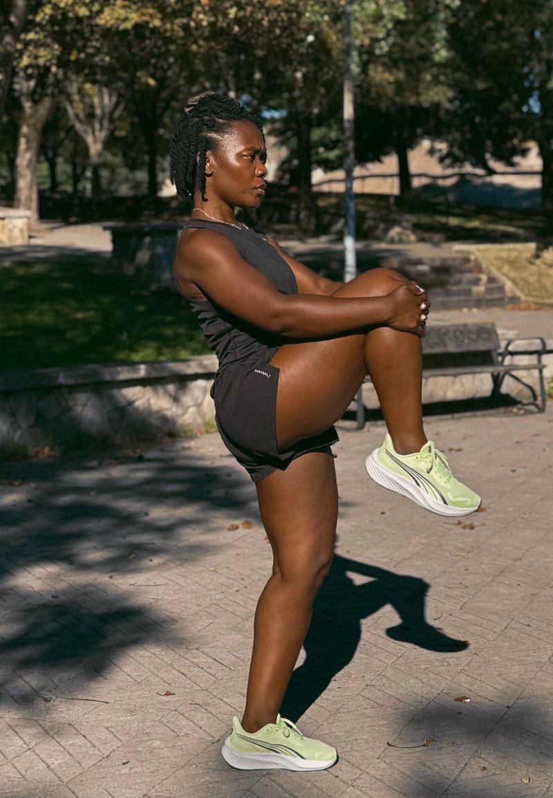 Woman in black athletic wear stretching one leg by pulling knee to chest outdoors on a sunny day in a park.