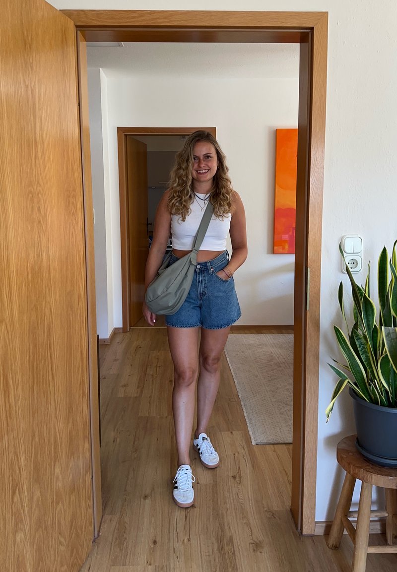 Ung kvinne med krøllete hår iført hvit topp og denimshorts som går gjennom en tredør i et lyst, moderne rom.