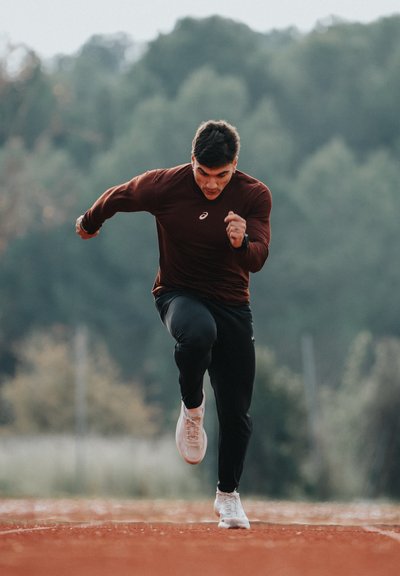 Hombre atlético con camiseta de manga larga color vino tinto y pantalones negros corriendo en pista roja. Zapatillas blancas, postura concentrada, fondo verde desenfocado.