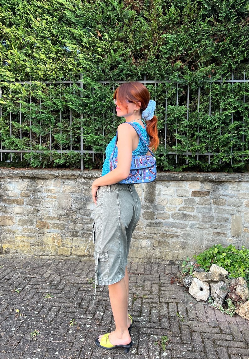 Μπλε καμπαρτίνα μπλούζα, γκρι cargo σορτς, κίτρινες ανοιχτές υποδήσεις. Μακριά κόκκινα μαλλιά με μπλε scrunchie, στέκεται μπροστά σε μια πέτρινη τοιχοποιία.