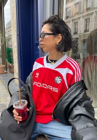 Rode Adidas-jersey met witte details en logo, gecombineerd met een zwarte oversized jas. Iemand houdt een transparante beker met een rietje vast.