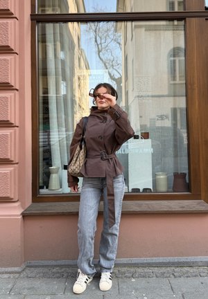 Mujer con chaqueta marrón con cinturón y vaqueros claros ajustándose las gafas de sol, de pie en la acera frente a un escaparate con cerámica en su interior.