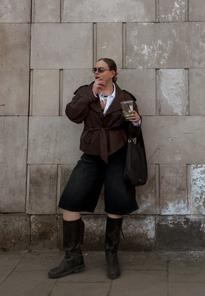 Femeie cu geacă închisă la culoare, ochelari de soare și cizme până la genunchi ține o băutură și stă în picioare în fața unui perete texturat de piatră, în aer liber.