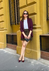 Bordeaux blazer over een witte T-shirt, een luipaardprint rok, een zwarte handtas en kastanjebruine puntige hakken. Achtergrond heeft een gele muur en een raam.