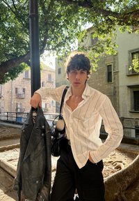 Witte gestreepte shirt, zwarte leren jas, zwarte cargobroek, en een zwarte tas. Buitenomgeving met bomen en stenen gebouwen op de achtergrond.