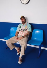 Gaiši zils un rozā krāsā blokota krekls ar grafisku printu, bēšas brīvas bikses un brūnas sporta kurpes uz zila grīdas pretī zilai sieniņai.