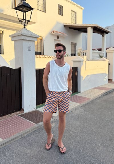 Top sin mangas blanco, pantalones cortos de estampado en naranja y azul con diseño de escamas de pescado, y chanclas negras. El escenario es una calle residencial.