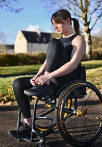 Fauteuil roulant noir avec des accents jaunes. Personne portant des leggings noirs et des chaussures, en train de faire ses lacets dans un parc, avec des arbres en arrière-plan.