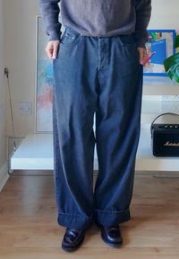 Széles szárú, sötét farmernadrág magas derékkal, ötpántos zsebes kialakítással és felhajtott szegéllyel, párosítva sötét bőrcipővel. Fa padló háttérrel.